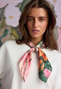 Hedvábný šátek s růžovými, oranžovými a zelenými květinovými vzory, pečlivě uvázaný kolem krku, kontrastující s oblečením světlejší barvy.