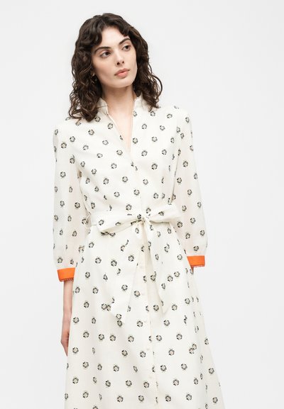 Femme portant une robe blanche à petits motifs floraux, avec des poignets bordés d’orange et une ceinture nouée à la taille, regardant sur le côté devant un fond uni.