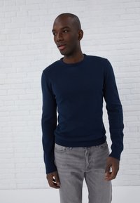 Marineblauw sweater van een zachte stof met een ronde hals en geribbelde manchetten. Draagt bij lichte grijze jeans, tegen een witte bakstenen achtergrond.