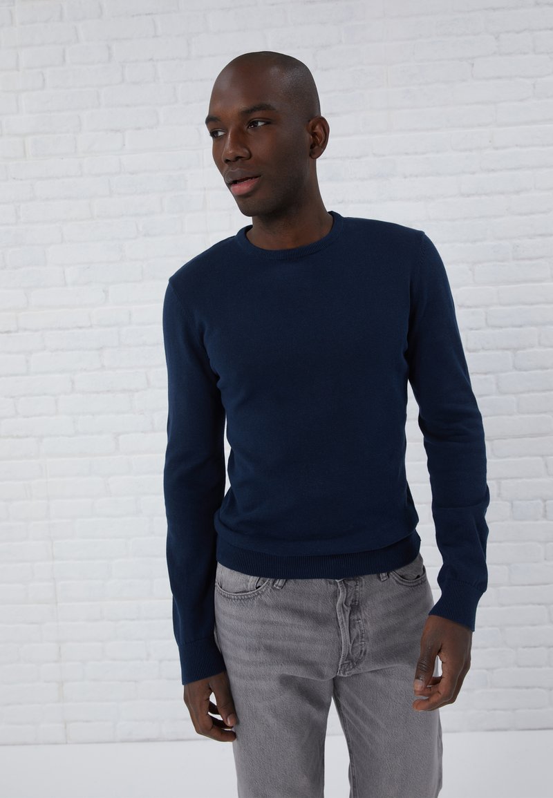 Marineblauw sweater van een zachte stof met een ronde hals en geribbelde manchetten. Draagt bij lichte grijze jeans, tegen een witte bakstenen achtergrond.