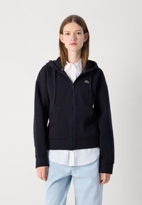 Sudadera con cremallera de color azul marino en tejido grueso, cuenta con una capucha con cordón y bolsillos delanteros, con un pequeño logo verde en el pecho.
