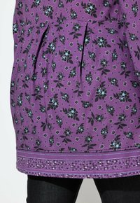 Fioletowa tunika w kwiaty z czarnymi i jasnoniebieskimi kwiatami, z plisami i dekoracyjnymi koralikami wzdłuż brzegu. Gładki bawełniany materiał.