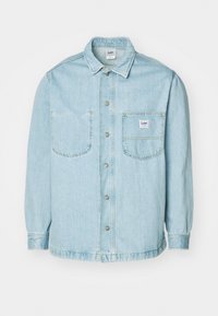 Veste en denim bleu clair avec manches longues, fermeture à boutons, deux poches poitrine et une étiquette cousue sur la poche gauche. Texture douce.