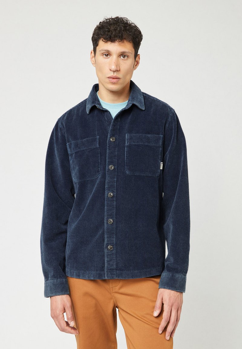 Camisa de pana azul marino con dos bolsillos en el pecho, cierre de botones y cuello. Mangas largas con detalles en los puños. Llevada sobre una camisa de un azul claro.