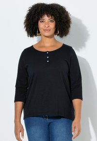 Haut noir à manches longues avec un col arrondi, doté d'une patte de boutonnage avec trois boutons. Texture douce ; coupe ample. Porté avec un jean bleu.