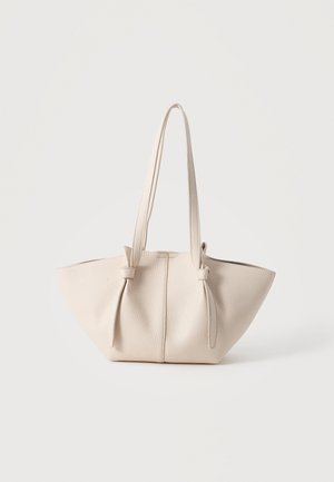 Beige strukturierte Ledertasche mit zwei langen Griffen, die oben in Knoten gebunden sind, vor einem schlichten hellen Hintergrund.