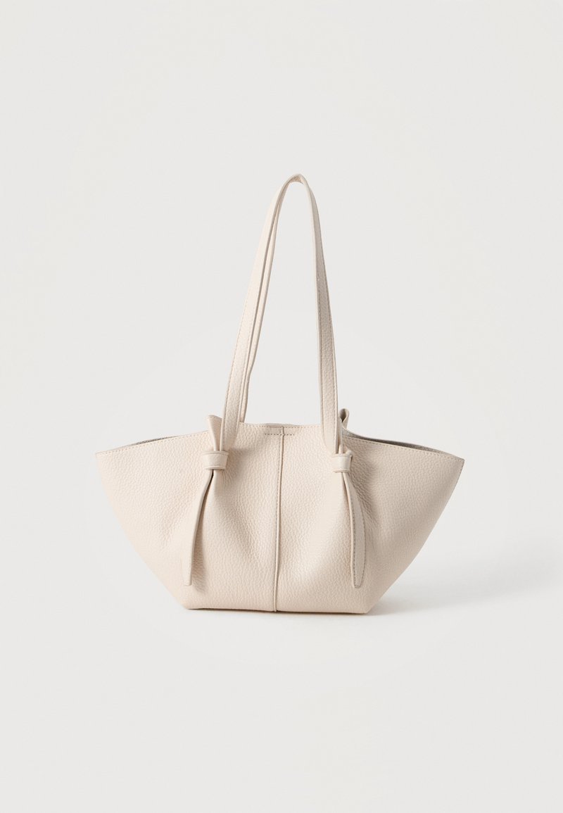 Sac cabas en cuir texturé beige avec deux longues poignées nouées près du haut, posé sur un fond clair uni.