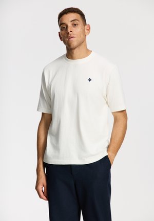 Mees, kes kannab valget lühikeste varrukatega T-särki, millel on rinnal väike tumesinine logo, ning tumedaid pükse, seisab lihtsa taustaga ettevalmistatud ruumis.