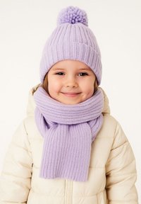 Lavendelfarbene Strickmütze mit einem Pompon, kombiniert mit einem passenden gerippten Schal, getragen über einer cremefarbenen Daunenjacke. Weiche Textur und gemütliches Design.