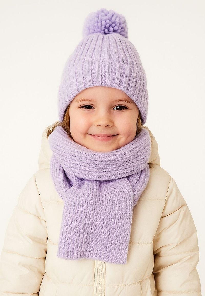 Lavendelfarbene Strickmütze mit einem Pompon, kombiniert mit einem passenden gerippten Schal, getragen über einer cremefarbenen Daunenjacke. Weiche Textur und gemütliches Design.