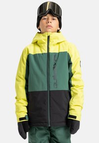 Junge Person trägt eine gelbe, grüne und schwarze Quiksilver-Winterjacke, schwarze Handschuhe, eine schwarze Mütze und Skibrille vor weißem Hintergrund.