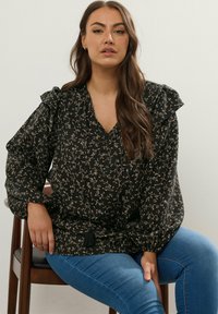 Blusa nera floreale con spalle a volant, dotata di scollatura a V e dettagli con frange, abbinata a jeans blu, mostrata mentre è seduta.