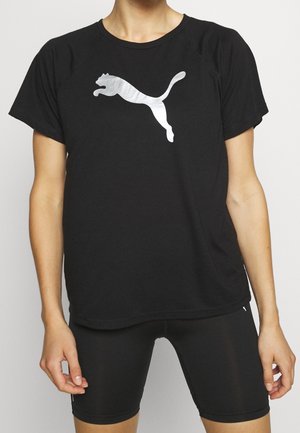 Osoba w czarnej koszulce Puma z srebrnym logo skaczącego kota i czarnych dopasowanych szortach kolarskich, stojąca na białym tle.