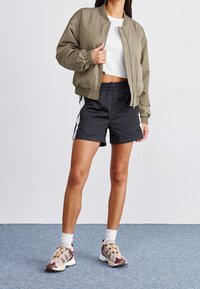 Olive green bomber jacket with elastic cuffs, white cropped tee, black athletic shorts with side stripes, and multicolored sneakers.