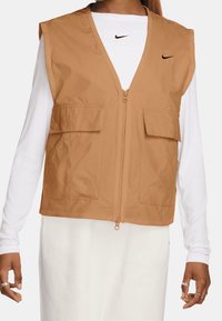 Béžová bezrukávová vesta se zipem, dvěma předními kapsami a nenápadným logem Nike na hrudi. Vyrobena z lehkého materiálu.