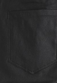 Zwarte denimstof heeft een achterzak met een chevronvormig ontwerp. Het oppervlak is glad met een lichte glans. De naden zijn zichtbaar langs de randen.