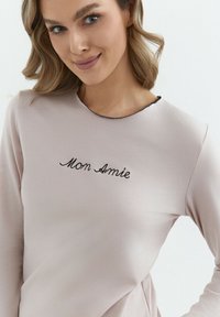 Jasnoróżowa bluzka z długim rękawem i okrągłym dekoltem, z czarnym haftowanym napisem "Mon Amie" na przodzie oraz subtelną fakturą.