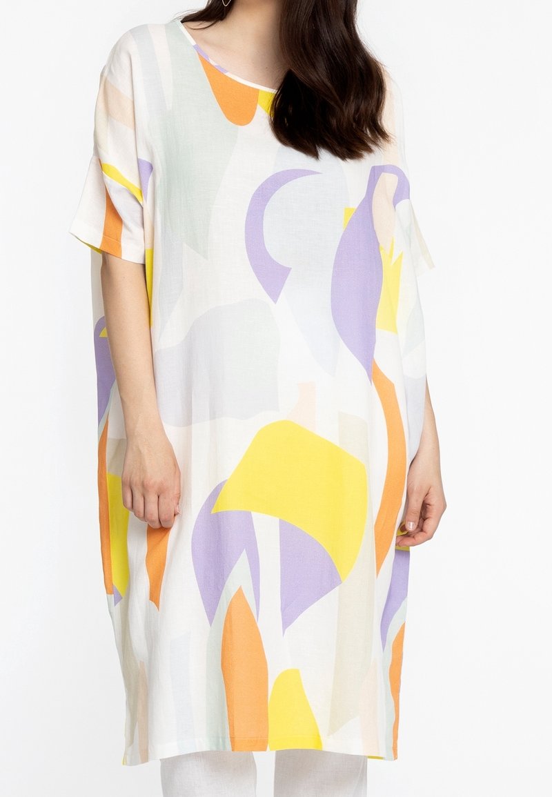 Femme portant une robe blanche ample avec des formes abstraites jaunes, orange et violettes, manches courtes et coupe droite.