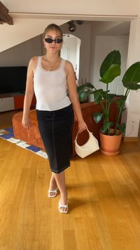 Bijela rebrasta majica bez rukava, crna traper suknja olovka sa šavovima, bijele sandale otvorenih prstiju i mala bijela torbica ispred pozadine s biljkama.