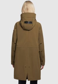 Manteau long khaki avec un col montant, doté d'un patch logo noir, de grandes poches et d'une fente arrière pour faciliter les mouvements. Texture lisse.