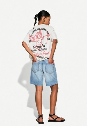 T-shirt bianca oversize con grafica rossa raffigurante un angelo, abbinata a pantaloni corti di jeans azzurri chiari e sandali neri.