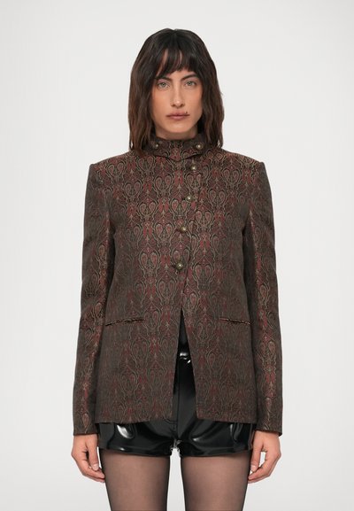 Femme portant un blazer à motifs, boutonné, avec un col haut, un short noir brillant et des collants noirs transparents, se tenant devant un fond uni.