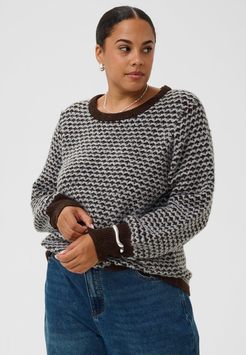 Femme aux cheveux foncés attachés, portant un pull marron et gris à motifs et un jean bleu, regardant vers la gauche.