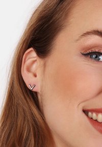 Silberfarbene Ohrstecker mit V-förmigem Design, flacher Oberfläche und minimalistischem Stil, die am Ohrläppchen getragen werden, vor einem neutralen Hintergrund.