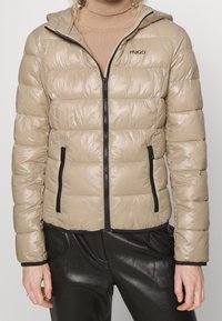 Beżowa puchowa kurtka z błyszczącym wykończeniem, zamek błyskawiczny i boczne kieszenie. Posiada kaptur i minimalne czarne logo.