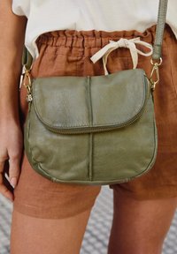 Olivgrüne Leder-Crossbody-Tasche mit einer runden Klappe, einem Reißverschluss-Frontfach und goldenen Hardware-Akzenten. Glatte Textur mit genähten Details.