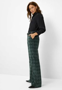 Pantaloni a zampa d'elefante verde scuro con motivo a trama, dotati di tasche laterali e cintura nera, abbinati a una camicia nera con colletto.