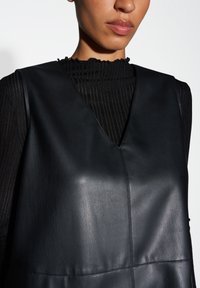 Zwarte leren vest met een V-hals ontwerp, met een gladde textuur en minimale stiksels. Daaronder een zwart ribbeltop.