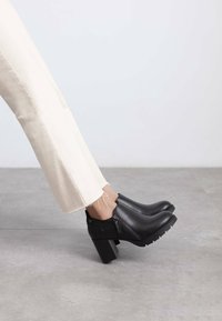 Botas de cuero negro hasta el tobillo con un tacón grueso, cremallera lateral y suela de goma texturizada, combinadas con pantalones cortos color crema.