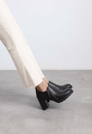 Botas de cuero negro hasta el tobillo con un tacón grueso, cremallera lateral y suela de goma texturizada, combinadas con pantalones cortos color crema.