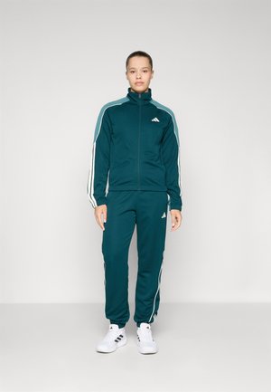 Teal trainingspak van zacht materiaal met een hoge kraag. Bevat witte strepen langs de mouwen en broekspijpen. Witte sneakers maken de look compleet.