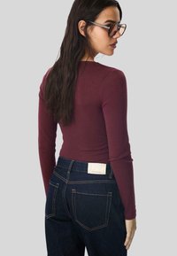 Langärmliges, kastanienbraunes Oberteil, kombiniert mit dunklen Jeans, die sichtbare Nähte und ein Markenetikett auf der Gesäßtasche aufweisen.