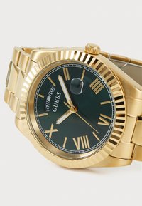 Montre dorée avec un cadran vert, des marqueurs en chiffres romains, un affichage de la date et une lunette texturée. Bracelet en métal avec des maillons polis.