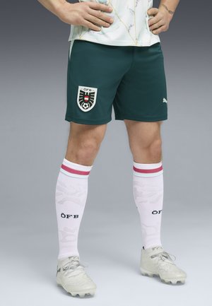 Atleta masculino vistiendo pantalones cortos verde oscuro con el escudo de fútbol ÖFB, tacos blancos Puma y calcetas blancas con rayas rojas y letras ÖFB.