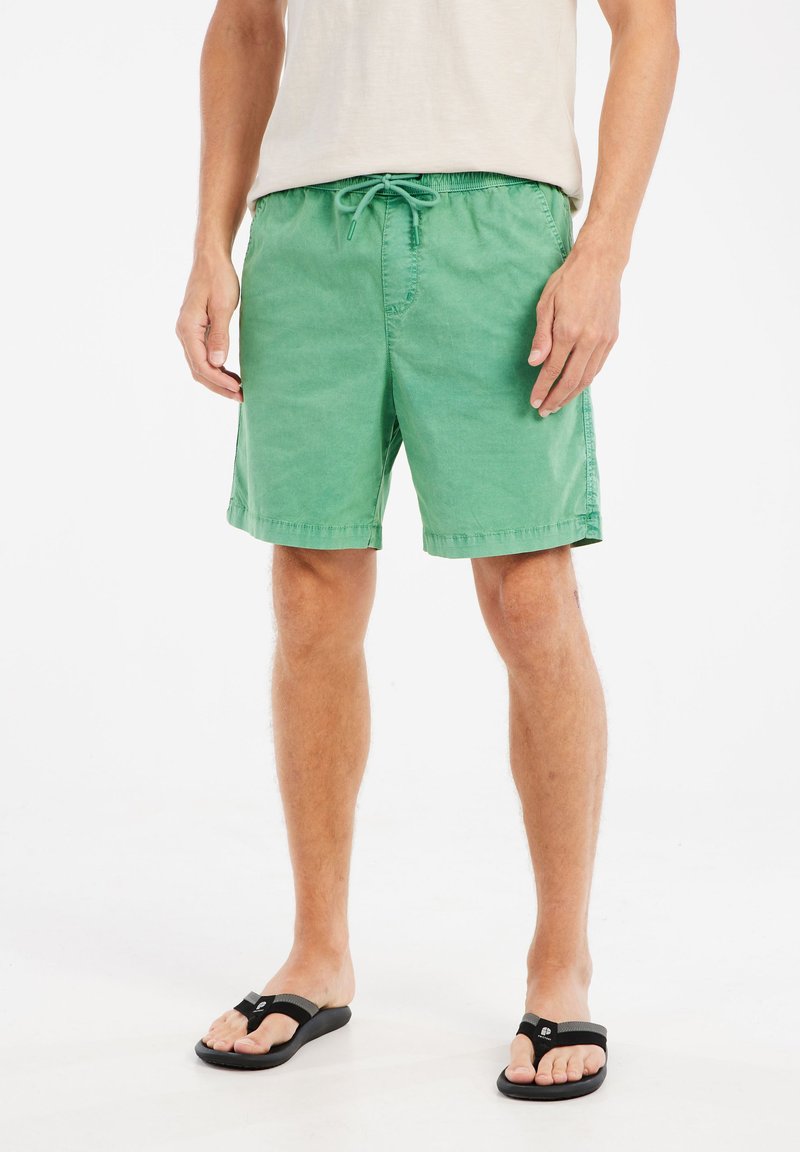 Groene katoenen zwemshorts met een elastische tailleband en drawstring, voorzien van zijzakken en een relaxte pasvorm, gecombineerd met zwarte teenslippers.