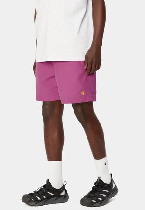 Bermudas moradas con una textura suave, que presentan un logo dorado en el costado, combinadas con zapatillas deportivas negras y calcetines blancos de caña corta.