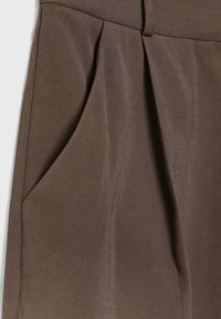 Gros plan sur un pantalon en tissu marron avec un design plissé et une poche latérale visible sur le côté gauche.
