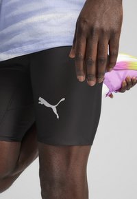 Sorte atletiske shorts med en glat overflade, prydet med et hvidt Puma-logo. En hånd holder en lilla og gul sko i baggrunden.