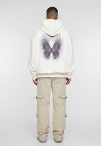 Sudadera blanca de gran tamaño con un gran gráfico de mariposa morada en la parte posterior. Combinada con pantalones cargo beige que tienen bolsillos laterales.