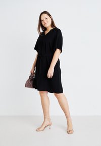 Robe noire à manches courtes avec un lien à la taille, présentant un décolleté en V. Tissu doux avec une coupe décontractée. Le modèle tient un petit sac rose et porte des sandales beiges.