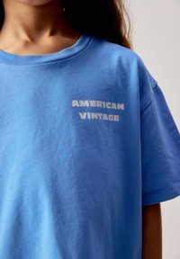 Enfant portant un T-shirt bleu clair avec le texte blanc "AMERICAN VINTAGE" sur la poitrine, sur un fond uni.
