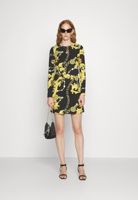 Vestido negro de manga larga con estampado barroco amarillo, de forma ajustada, combinado con gafas de sol negras chunky y un pequeño bolso negro.