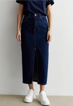 Frau trägt einen dunkelblauen Midi-Jeansrock mit einem Frontschlitz, kombiniert mit weißen Turnschuhen und einem dunklen, kurzärmligen Oberteil vor einem schlichten Hintergrund.