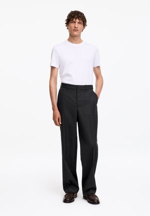 Man met krullend haar, gekleed in een wit kortemouwenhemd, donkergrijze broek en donkerbruine schoenen, staand met één hand in de zak.