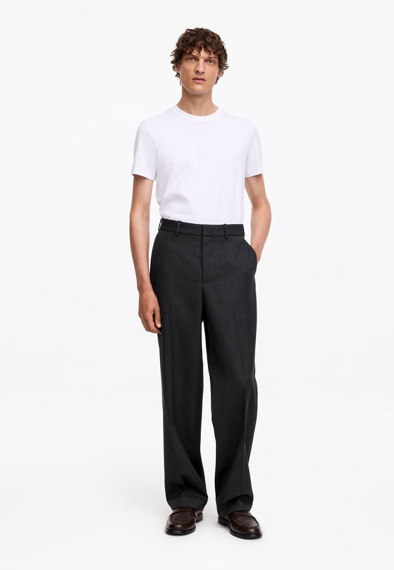 Homme aux cheveux bouclés portant une chemise blanche à manches courtes, un pantalon gris foncé et des chaussures marron foncé, debout avec une main dans la poche.
