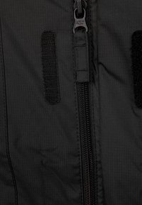 Svart tyg med en vertikal Dragkedja i mitten och två vertikala kardborreband på vardera sidan av dragkedjan.